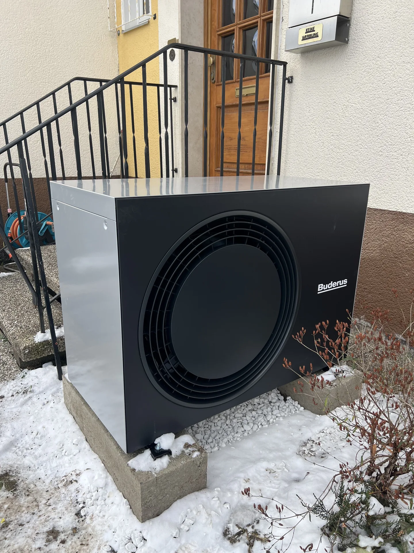 Wärmepumpen-Außeneinheit auf Betonsockel vor einem Hauseingang im Winter